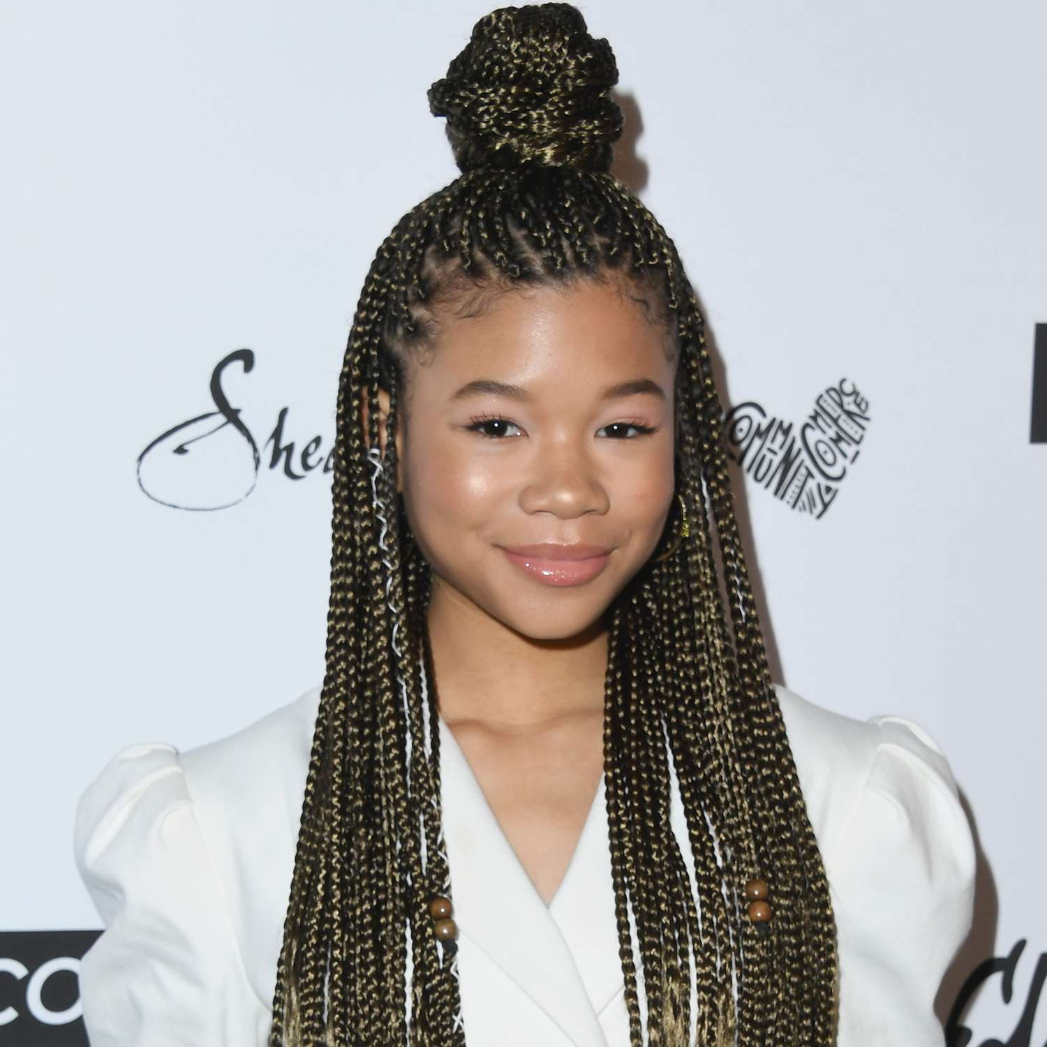 zoomed in picture of Storm Reid looking at camera with long braided hair, and top section piled into twisted braided bun on top of head