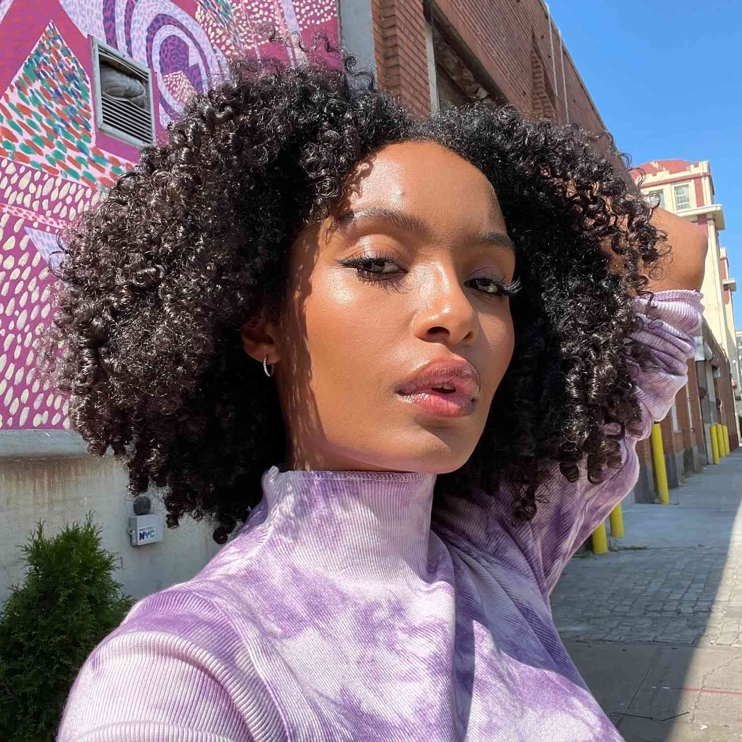 Yara Shahidi with a cropped, curly hairstyle, radiant skin, and a purple marbled turtleneck