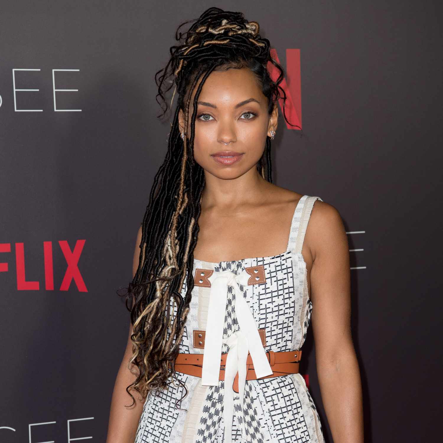 Logan Browning looking at camera with long braided hair, a few pieces left out as face-framers with the top half pulled back into a bun pile on top of head