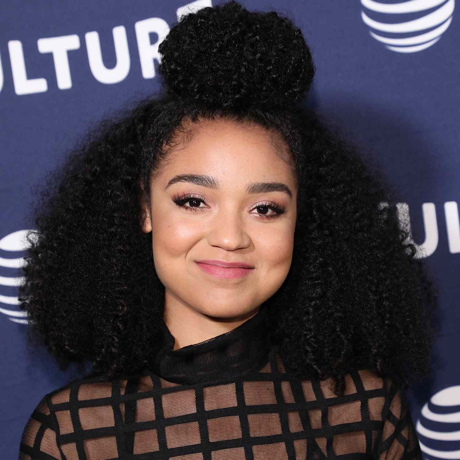 Aisha Dee smiling at camera, zoomed in picture showing off her shoulder length curls with top crown section pulled into large bun on top of head