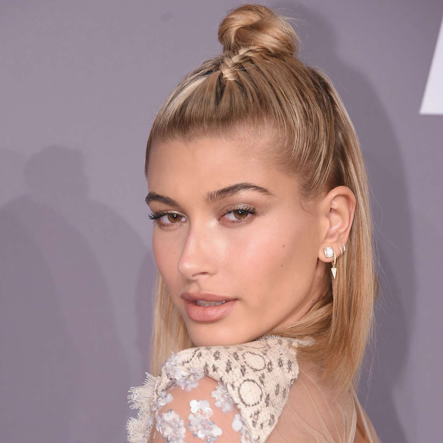 Hailey Bieber looking over shoulder at camera, zoomed in on face with short blonde straight bob and top section pulled back tightly with braid going back into a twisted bun on top of head