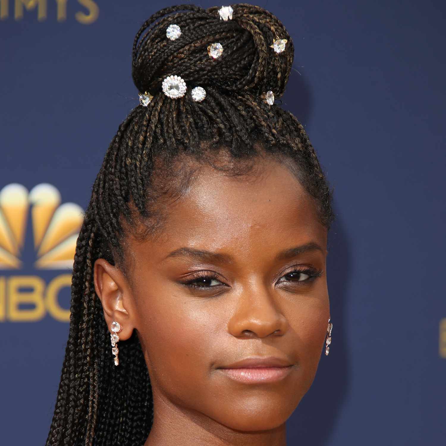 Letitia Wright looking at camera, zoomed in on hair in braids and pulled up into a large bun on top of head with crystal embellishments