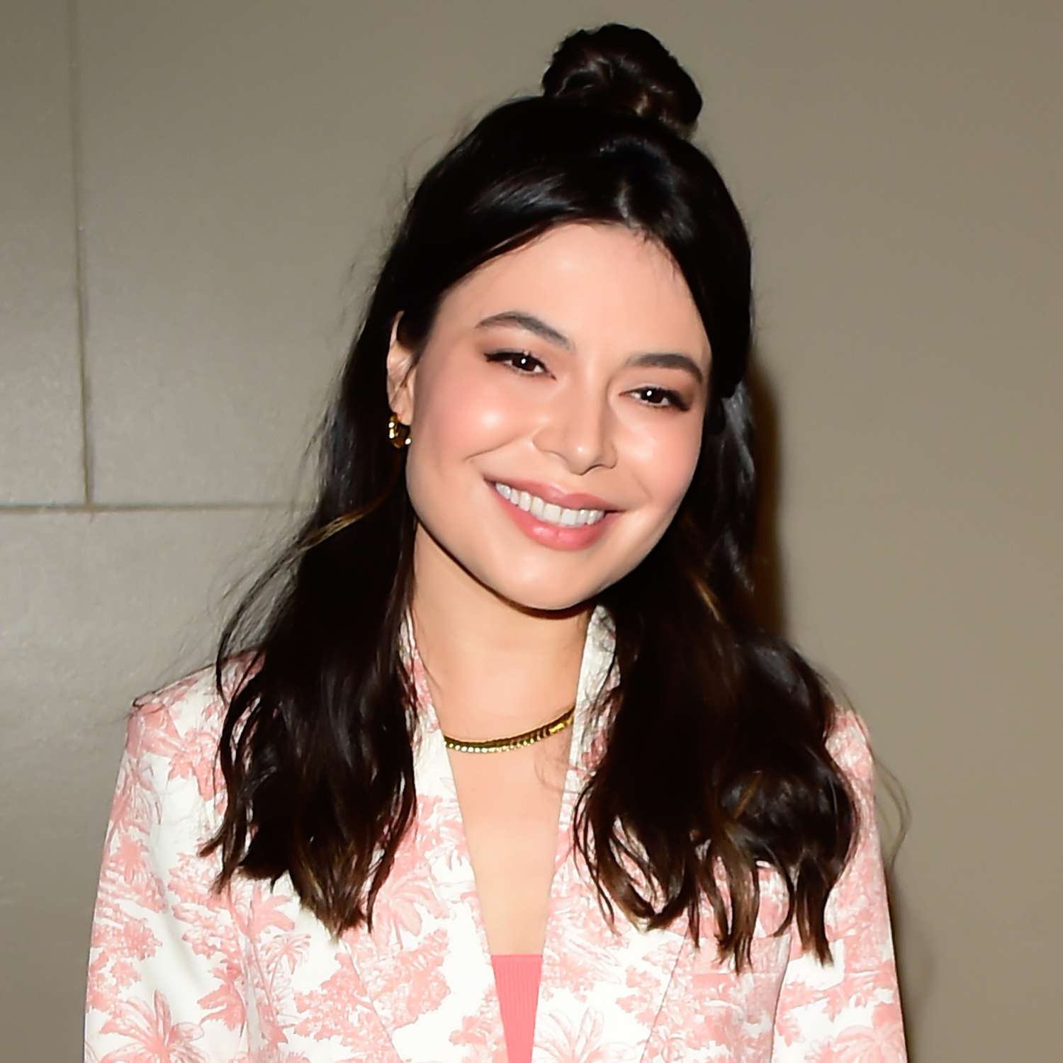 zoomed in picture of Miranda Cosgrove smiling at camera with shiny black waves, bangs left out, and top section of hair lightly pulled back into messy bun at the back top of the head