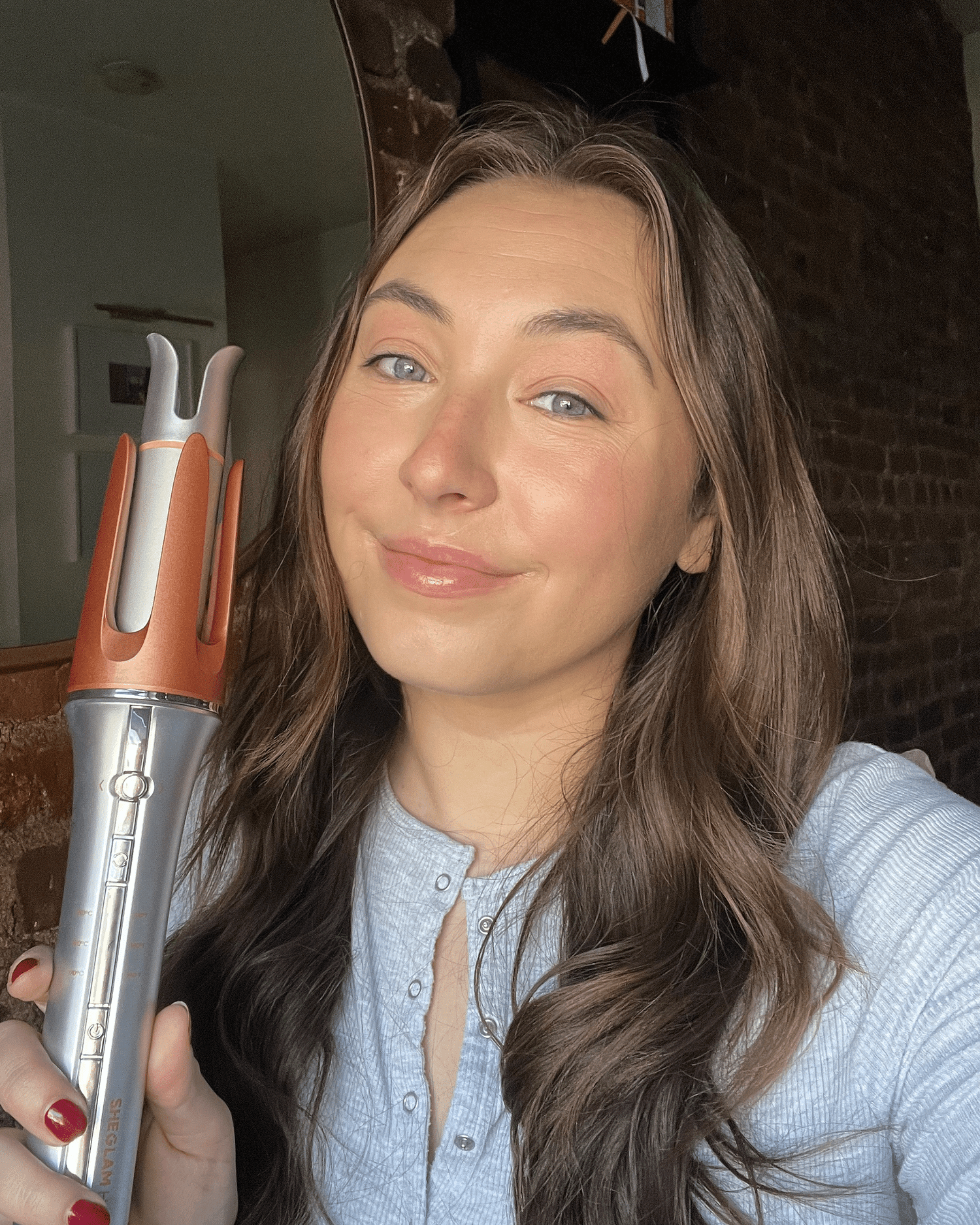 Girl with long curled brown hair holding the orange and silver SHEGLAM hair tool